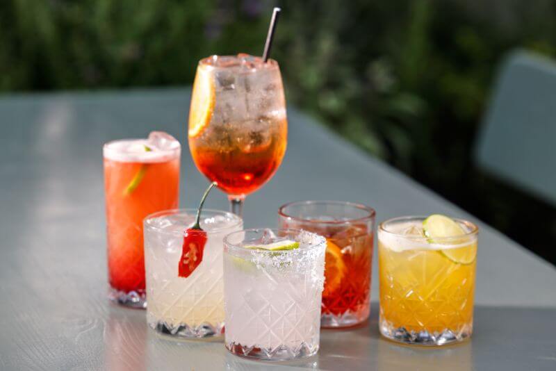 Six different cocktails are aligned on a table outdoors in the sunshine.