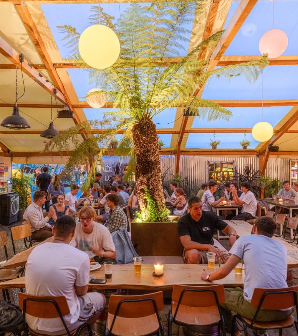 Peckham Arches interior at dusk, decorated with colourful globe lights in orange, pink and red, tropical ferns, and lush green plant walls. Guests socialise at tables beneath a clear roof.