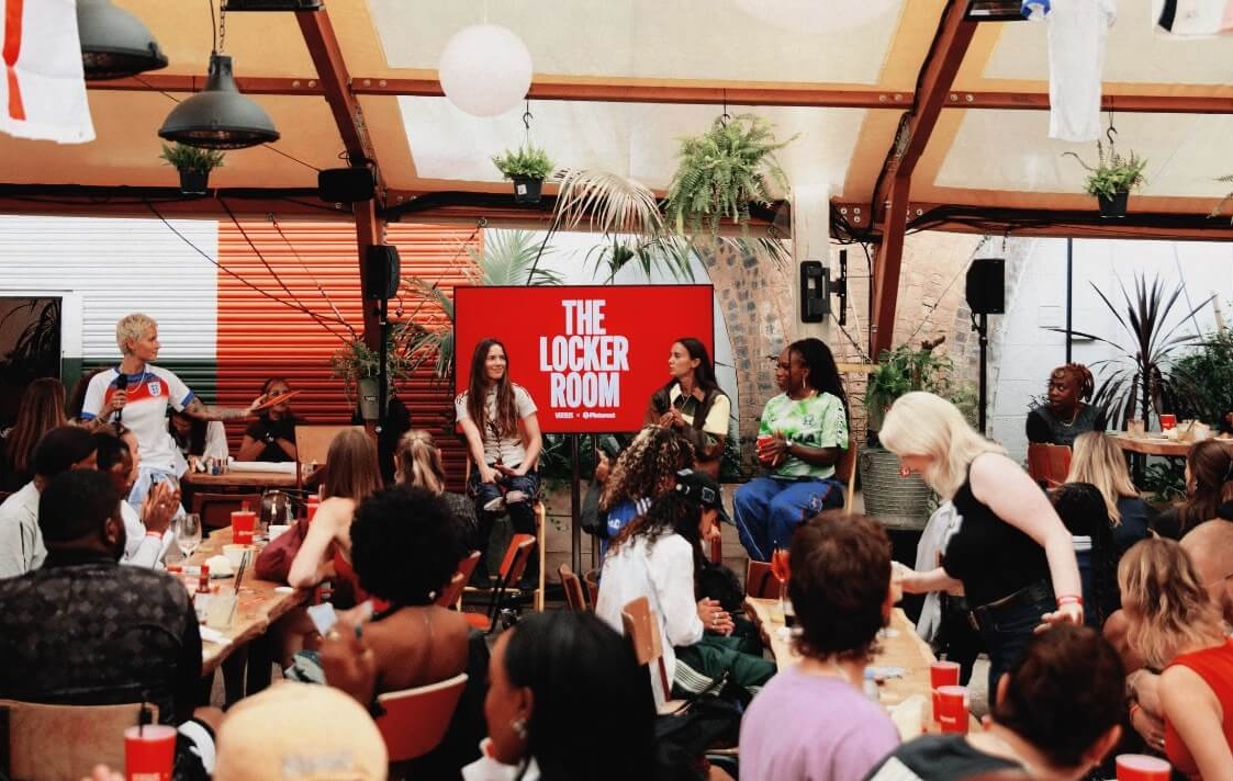 Peckham Arches interior at night, decorated with colourful globe lights in orange, pink and red, and tropical ferns. Guests socialise at tables beneath a clear roof.